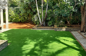 Lush garden with artificial grass, wooden deck, and tropical trees.